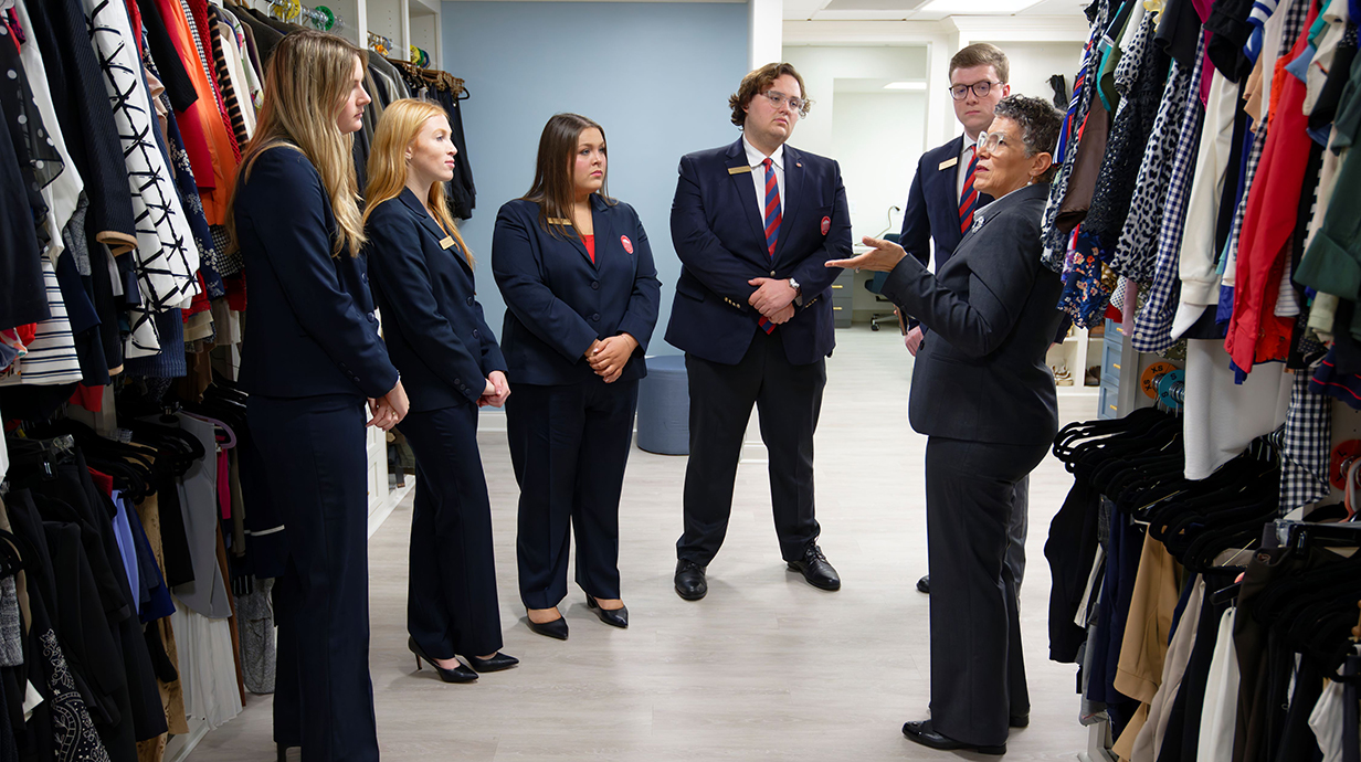 A woman talks to a group of young people, all dressed in business attire, as they stand between racks of clothing.