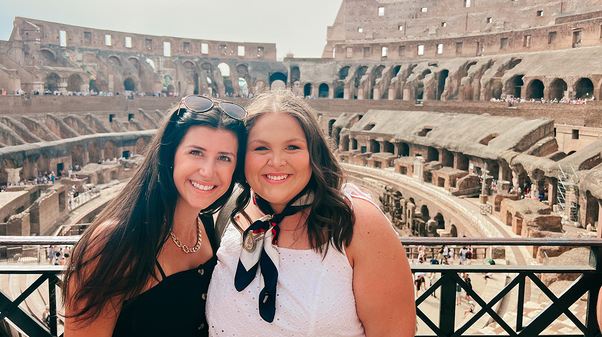 Two young women stand inside the Colosseum in Rome on a sunny day.