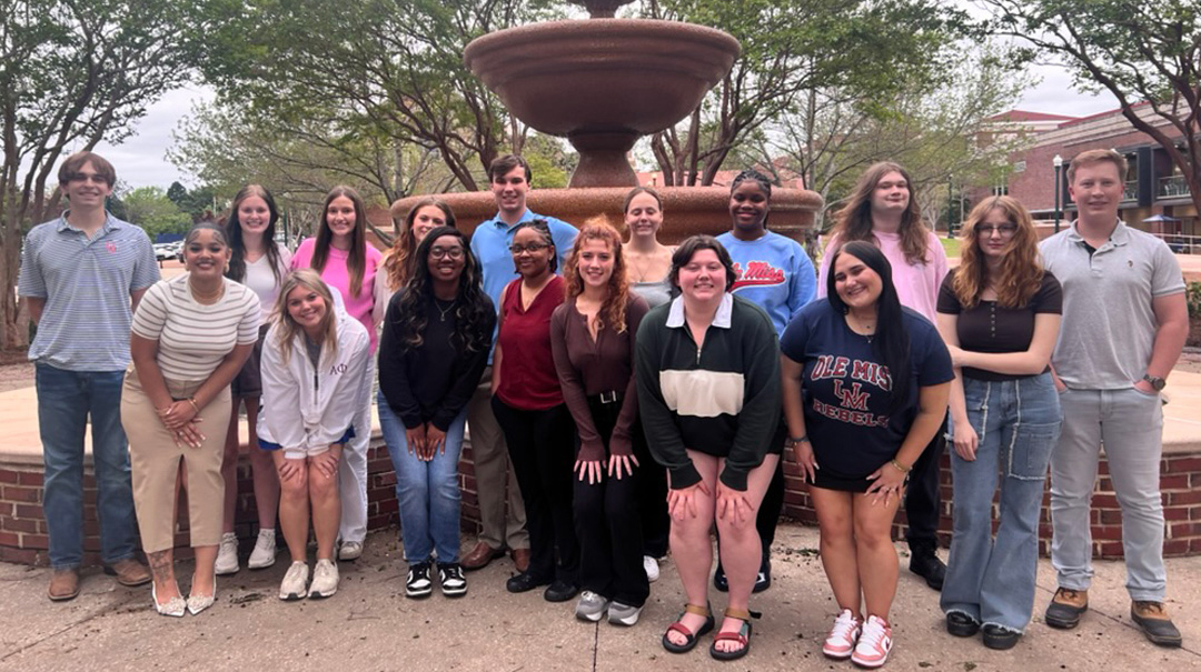 A group of young people stands in front of a fountain surrounded by trees.