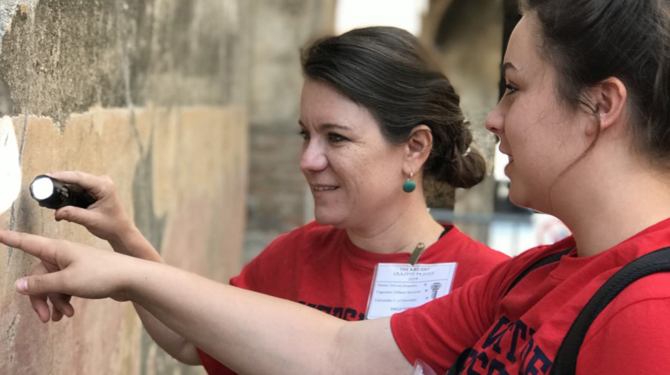 Two women waering red T-shirts use a flashlight to examine markings on a stone wall.