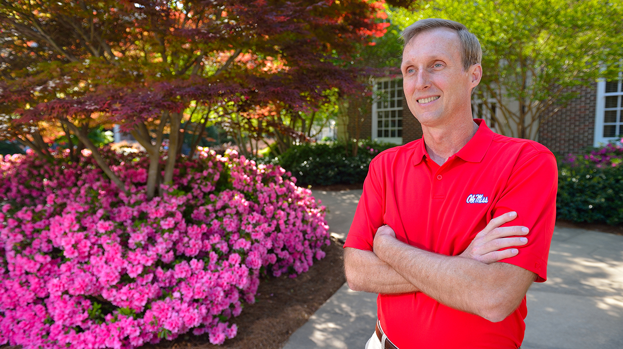 A man wearing a red shirt smiles as he stands in front of vibrant azaleas in bloom.