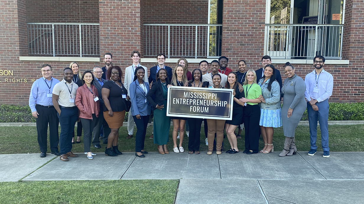 A large group of people poses outside a building while holding a sign that reads 'Mississippi Entrepreneurship Forum.'