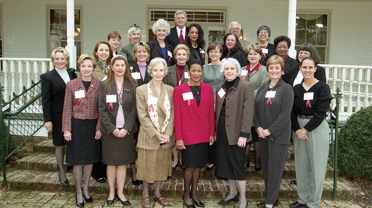 A large group of women and two men pose for a photo on steps leading to a white building.