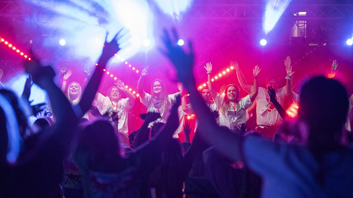 A group of young women dance on a stage lit by colorful spotlights as a crowd cheers and waves.