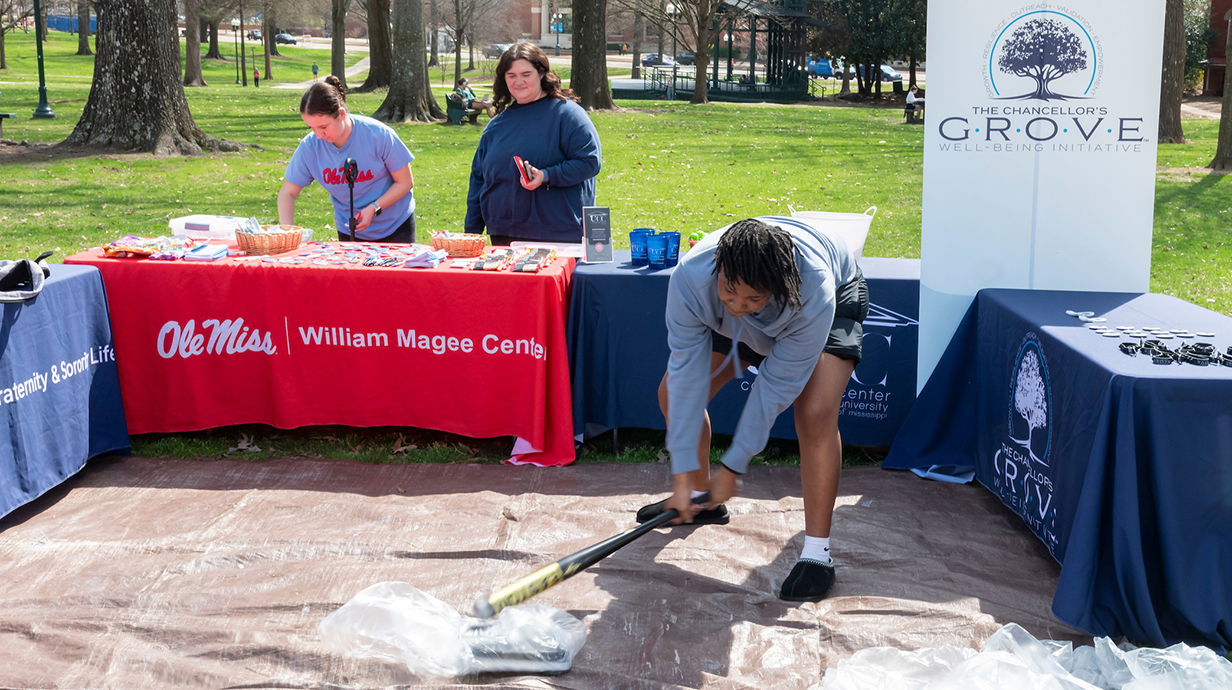 A young woman uses a baseball bat to smash a bathroom scale in a booth in an outdoor park.