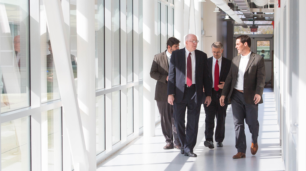 Four men in suits talk as they walk down a hallway lined with full-length windows on a sunny day.