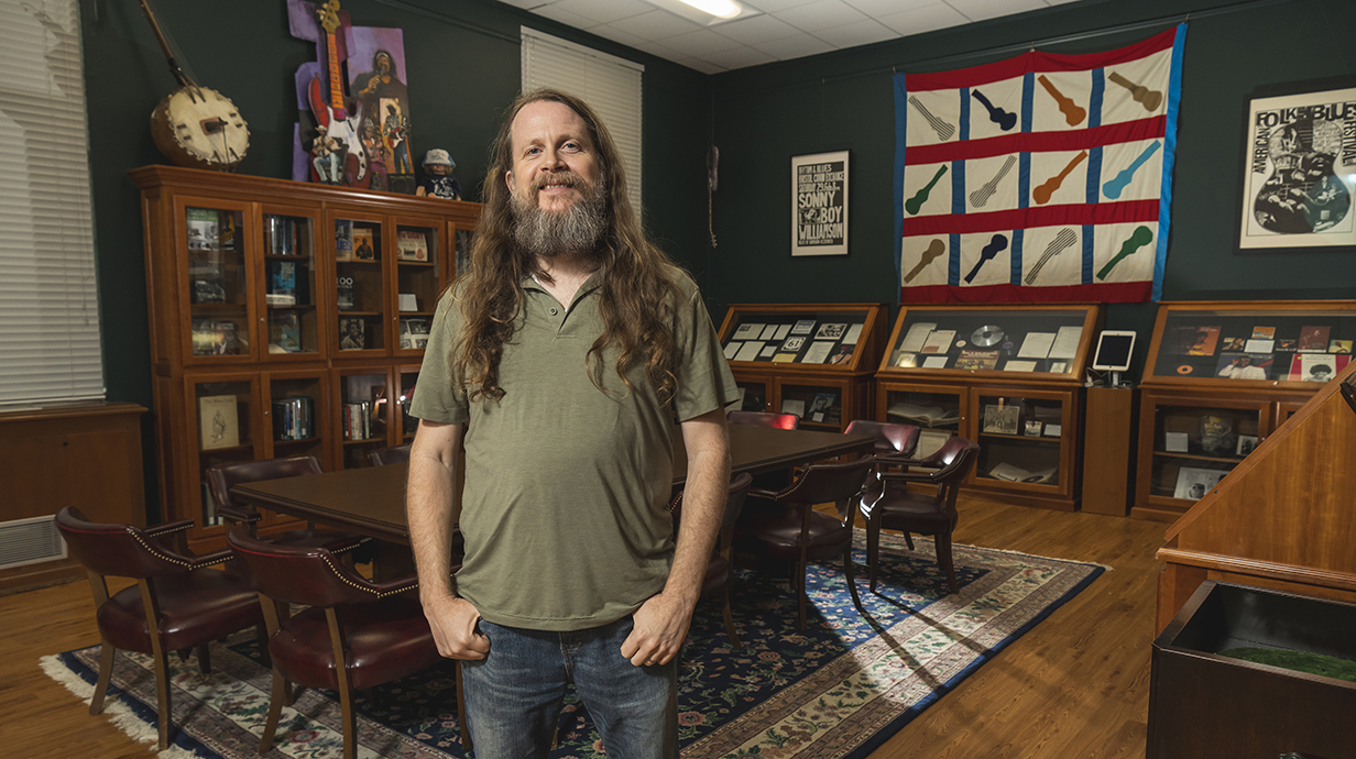 A bearded man with long hair stands in a library room lined with display cases full of vinyl records, photographs and other memorabilia. Posters and other artwork line frame the display cases.