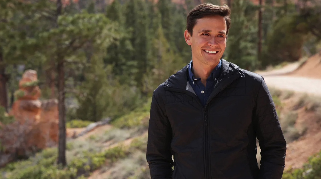 A young man wearing a dark jacket stands on a hillside in front of a wooded area.