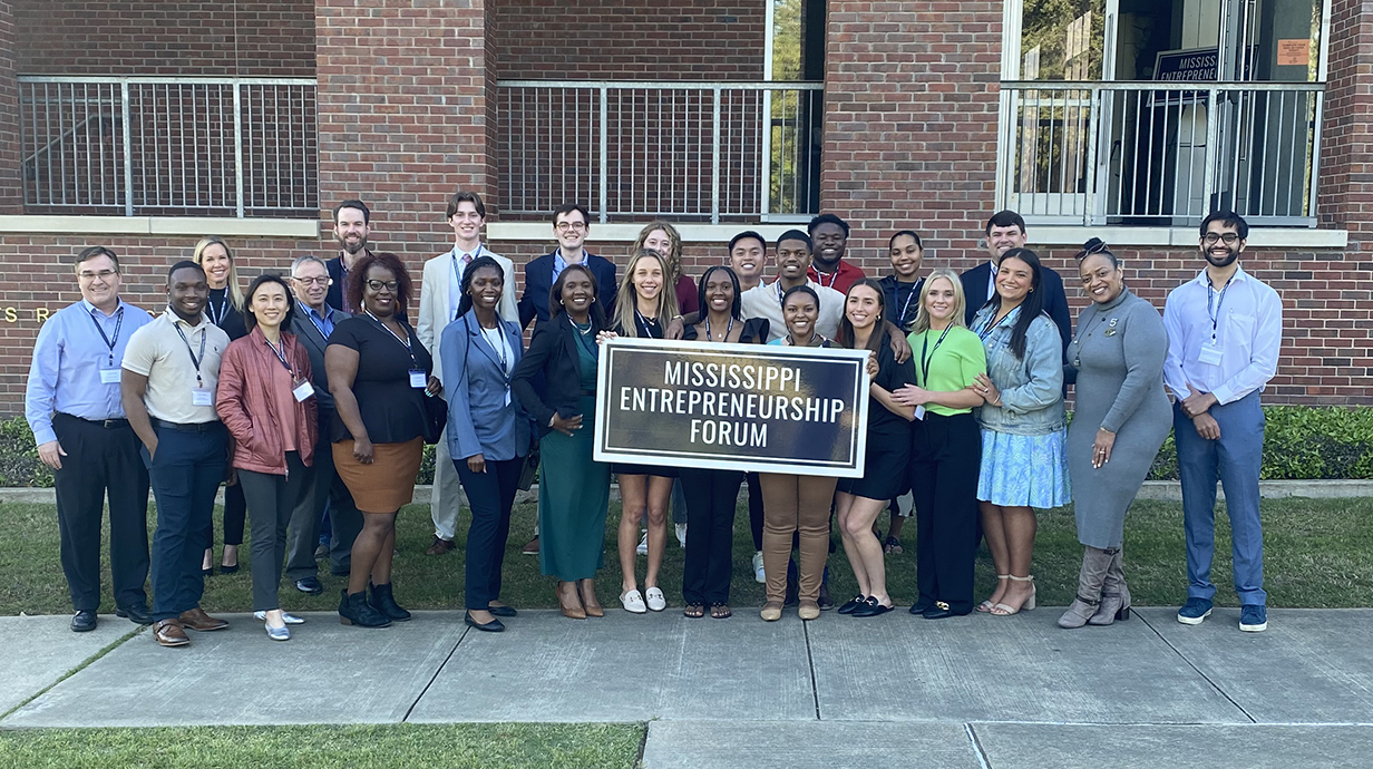 A large group of people stands outside a red brick building holding a sign that reads 'Mississippi Entrepreneurship Foruim."