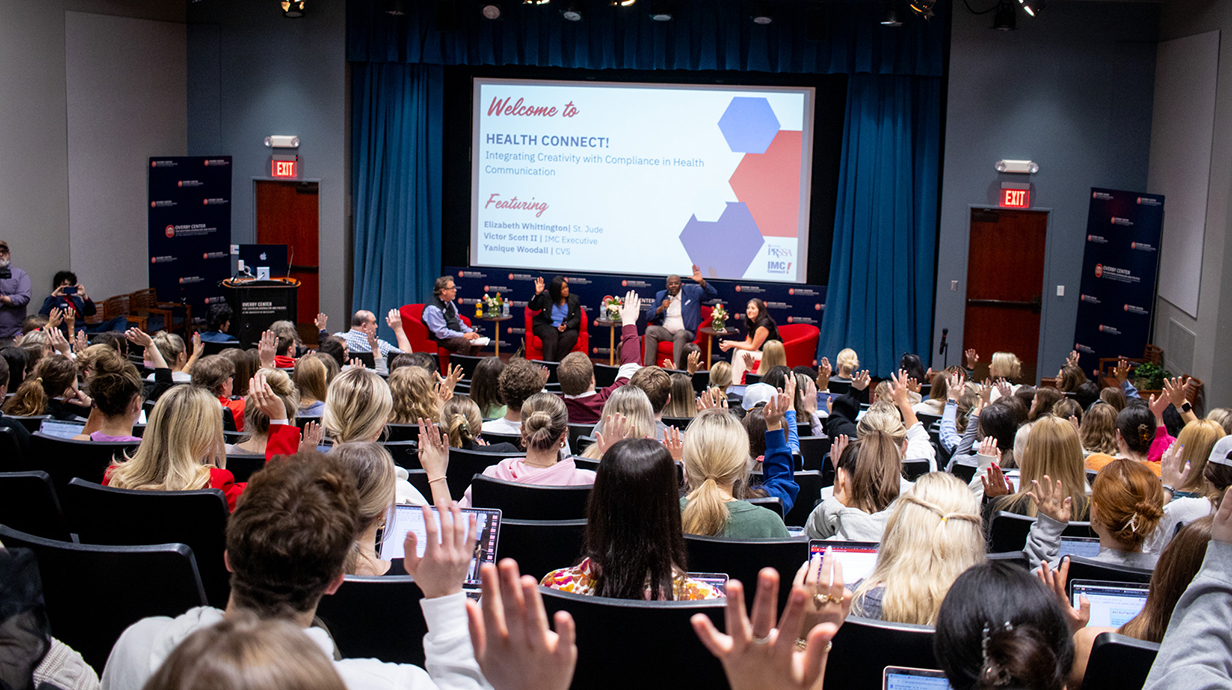 A crowd of people sitting in an auditorium raise their hands in response to four people sitting on a stage in the background.