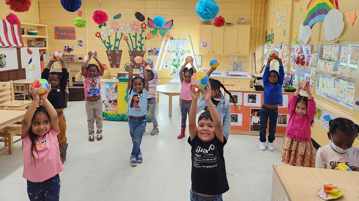 A group of young children stand in a bright yellow classroom holding inflatable balls over their heads.