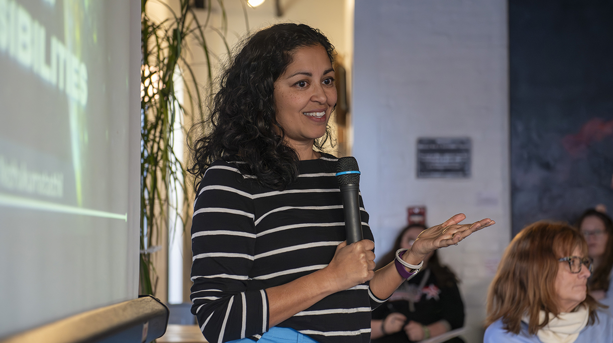 A woman holding a microphone stands in front of a projection screen and talks to a group of people.