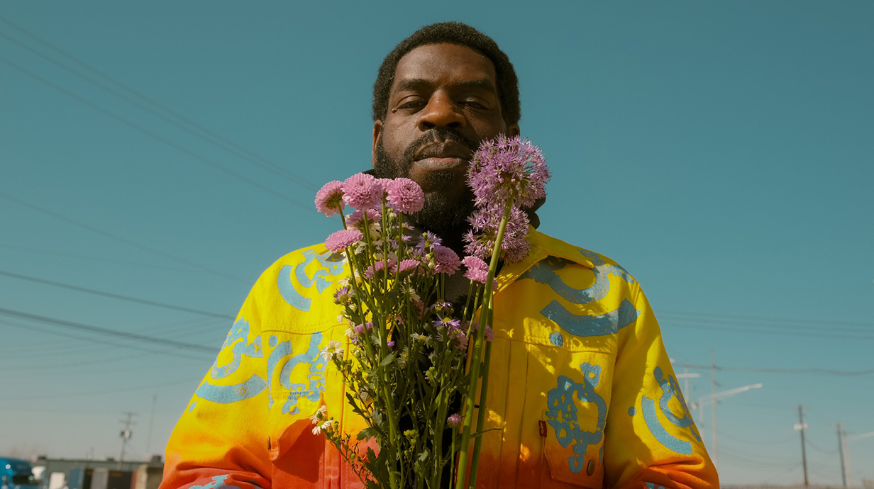 A man wearing a bright red-and-yellow shirt stands outdoors holding a bunch of wildflowers.