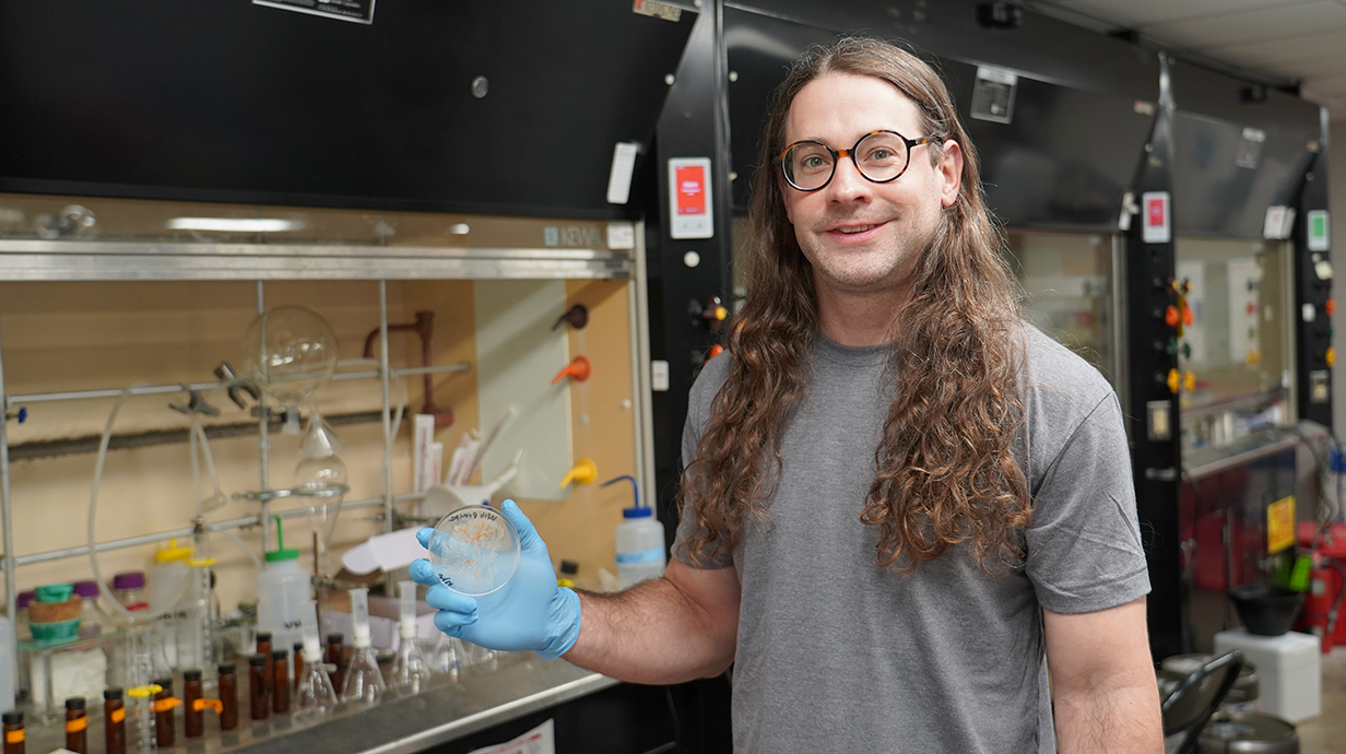 A man wearing glasses and blue gloves holds a petri dish containing a bacterial culture in a laboratory.