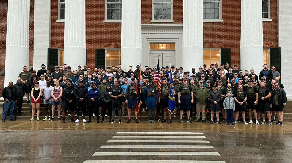 A large group of people gather on the sidewalk and steps in front of a large columned building on a rainy morning.