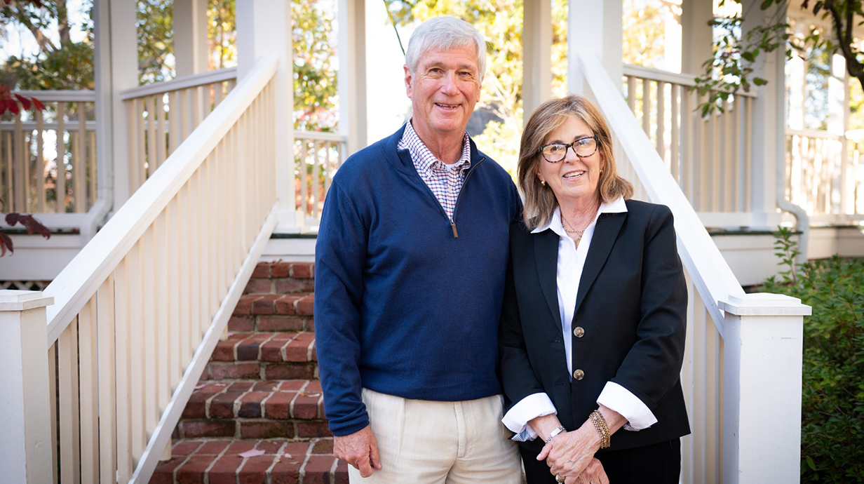 A man and woman pose for a photo standing on white steps leading up to a covered porch.