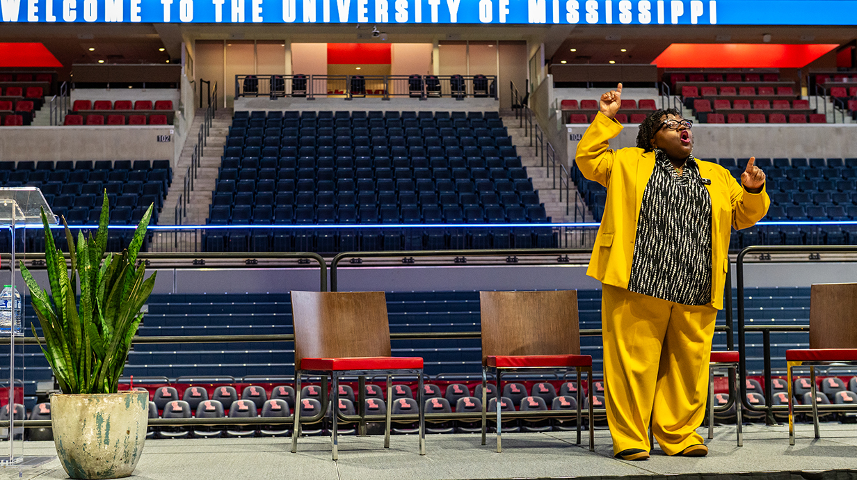 A woman wearing a bright yellow coat and pants talks from a stage.