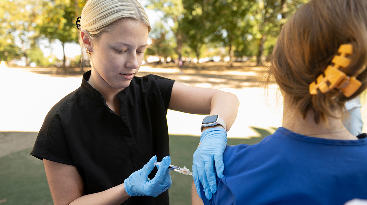 A young woman wearing blue gloves gives another young woman an injection in her upper arm.