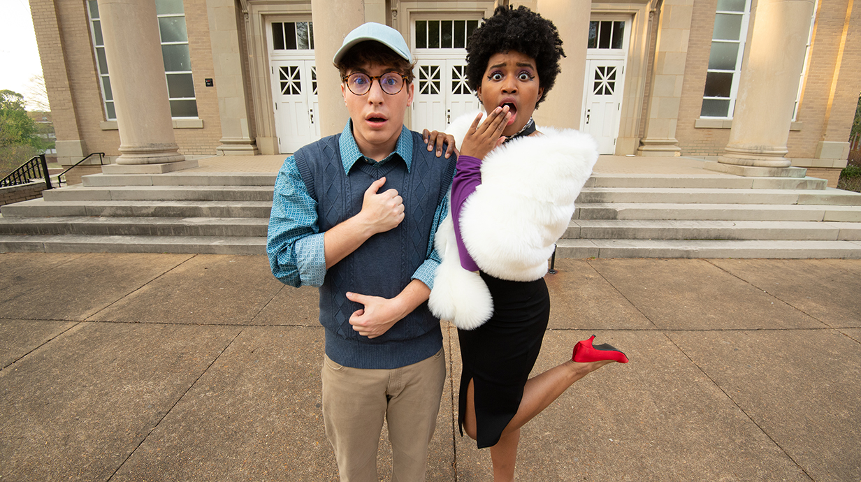 A young man and young woman, both with startled expressions, stand in front of a  stone theater.