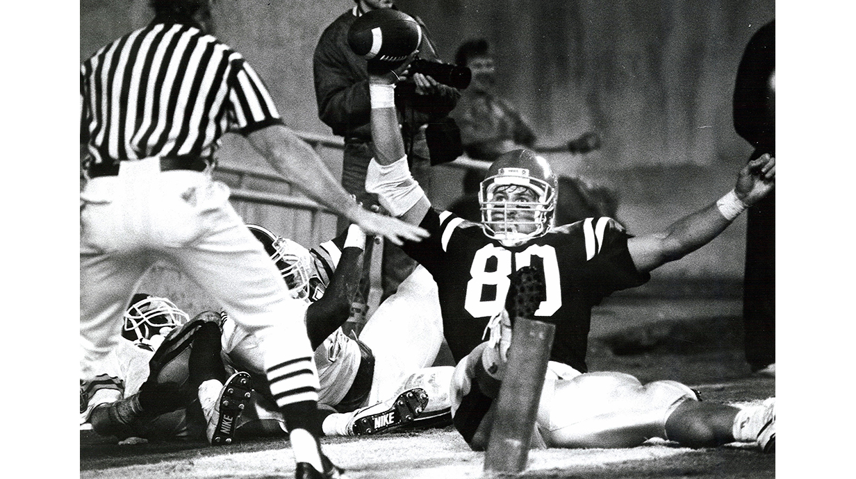 A football player sitting on the turf during a game holds up a football as a referee signals.