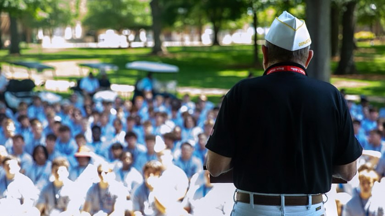 A man with his back to the camera addresses a large group of young men from an outdoor stage in a wooded park.