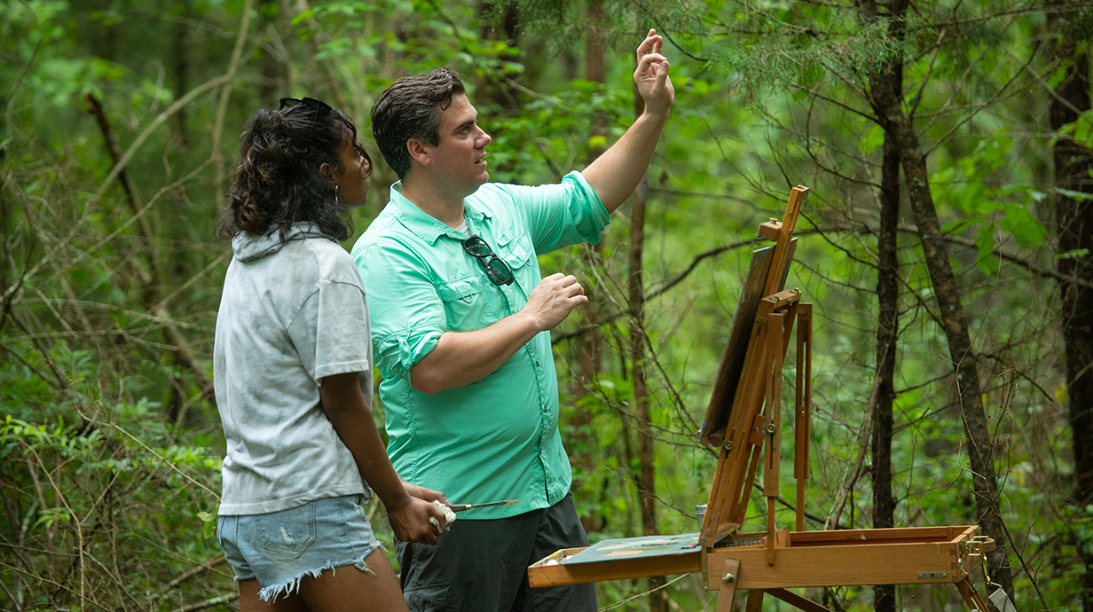 A man standing at an artist's easel gestures into the distance while a young woman holding a paintbrush watches in a tree-lined area.