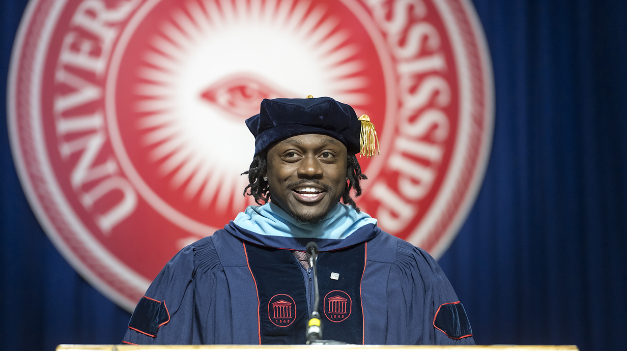 A man dressed in a graduation robe and cap speaks at a ppodium in front of a red University of Mississippi seal.