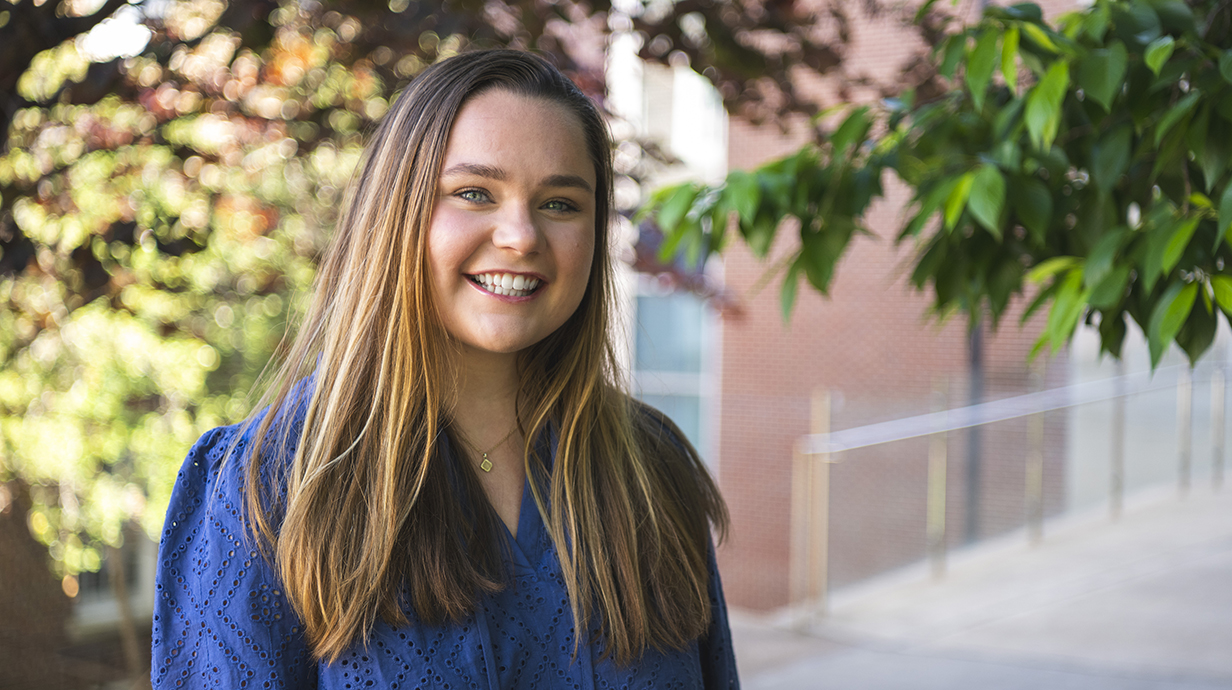 A young woman wearing a blue blouse stands underneath trees in front of a red brick building.