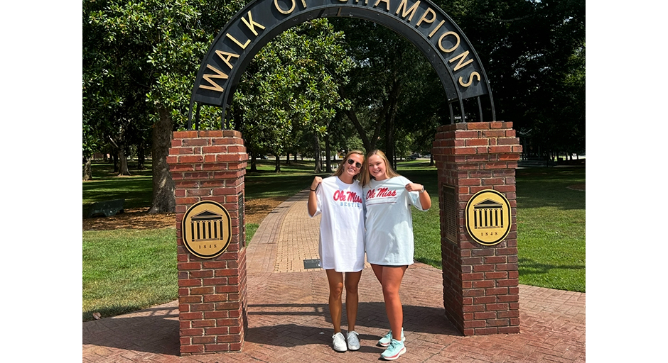 Two young women, both wearing Ole Miss T-shirts, stand underneath an arch reading 'Walk of Champions' on a sunny day.