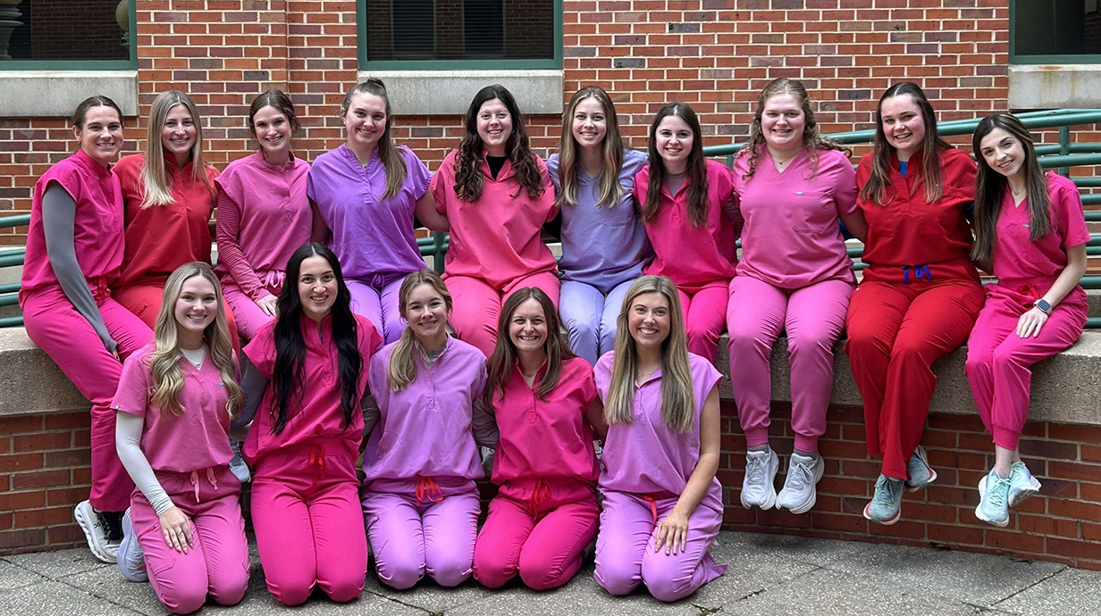 A large group of young women, all wearing red and pink scrubs, pose for a photo outside a brick building.