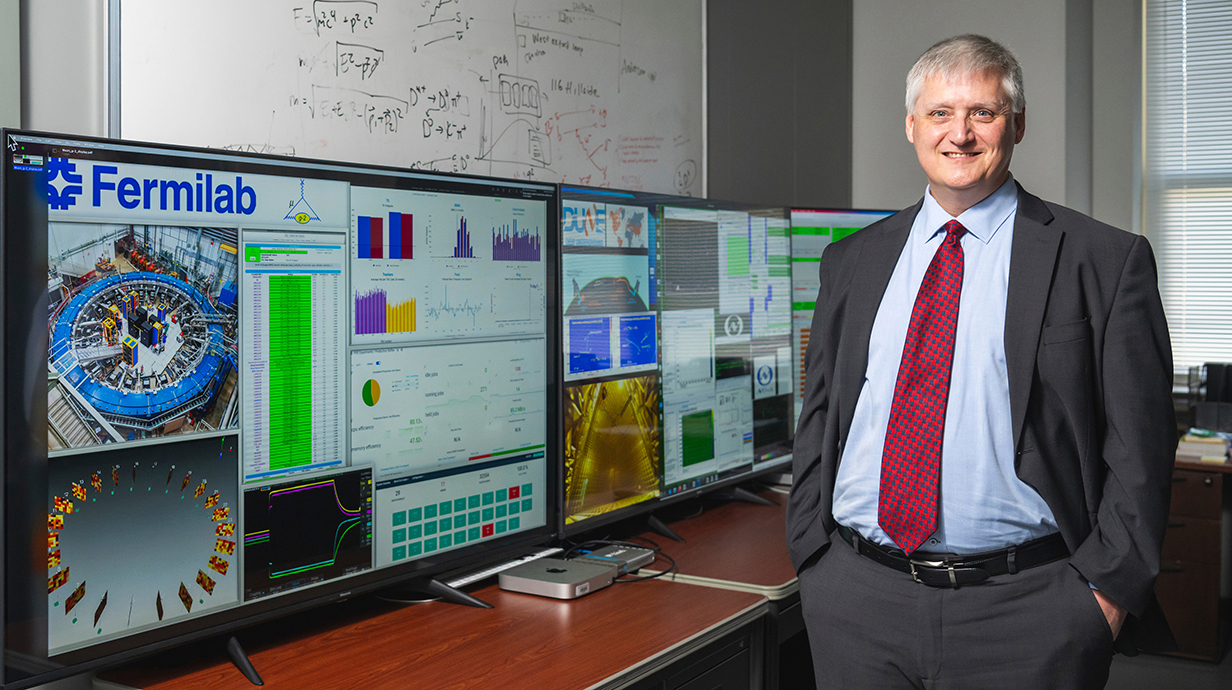 A man wearing a suit stands by a research display for Fermilab.
