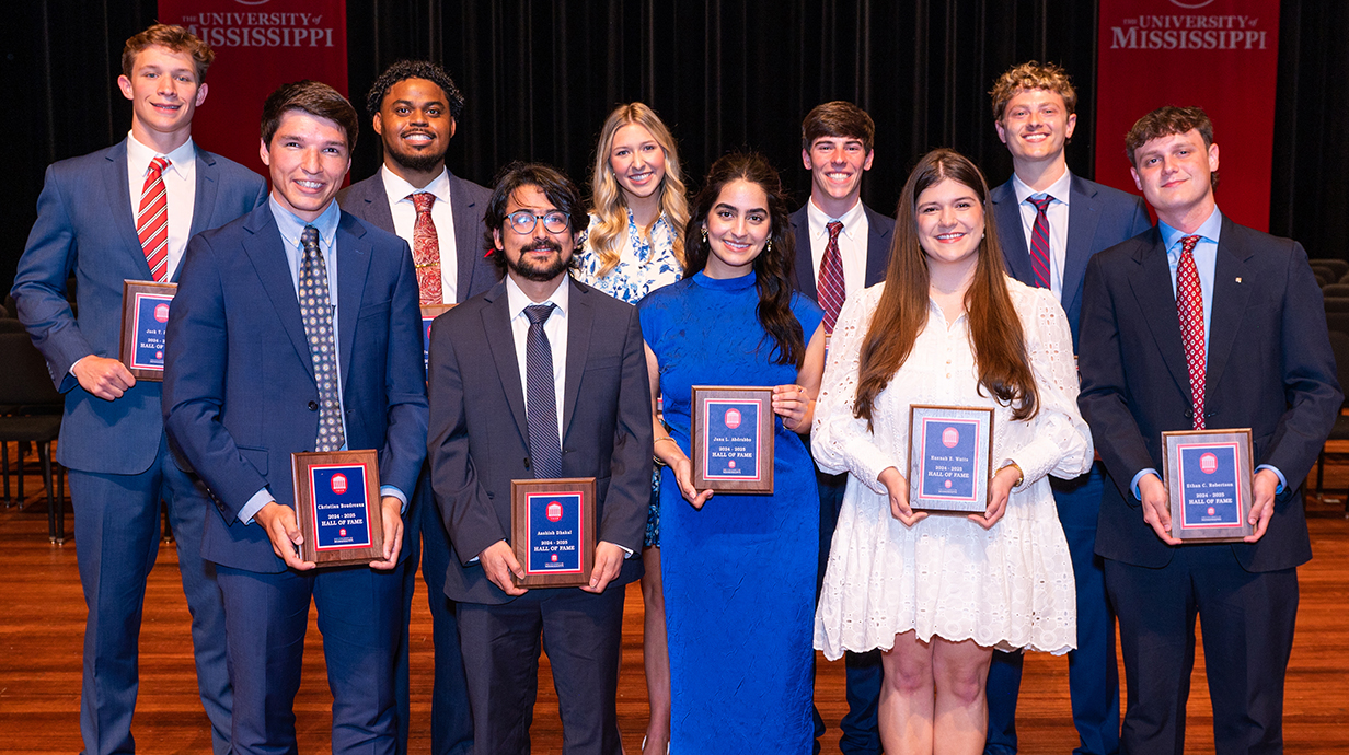 A group of young people in business attire, all holding award plaques, stand on a stage.