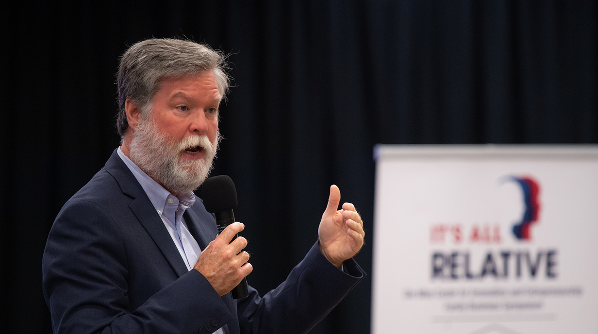 A man wearing a suit speaks into a microphone in front of a projection screen.