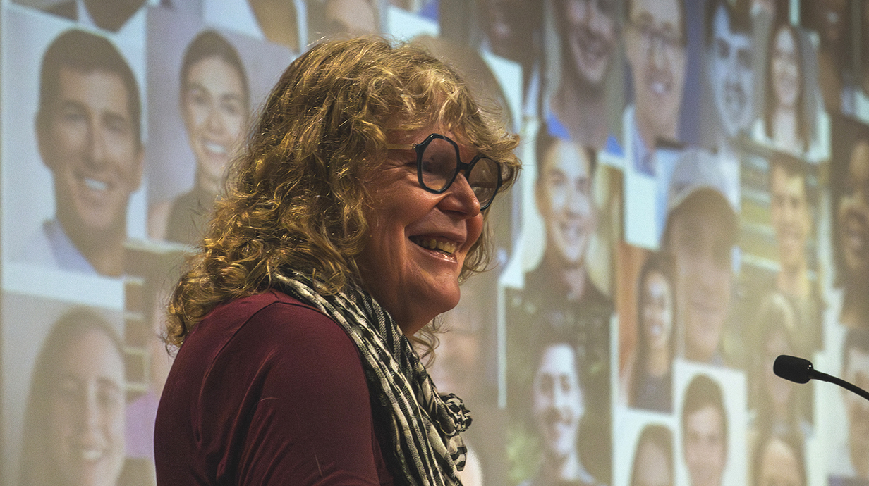 A woman stands in front of a projection screen displaying faces of dozens of young people.