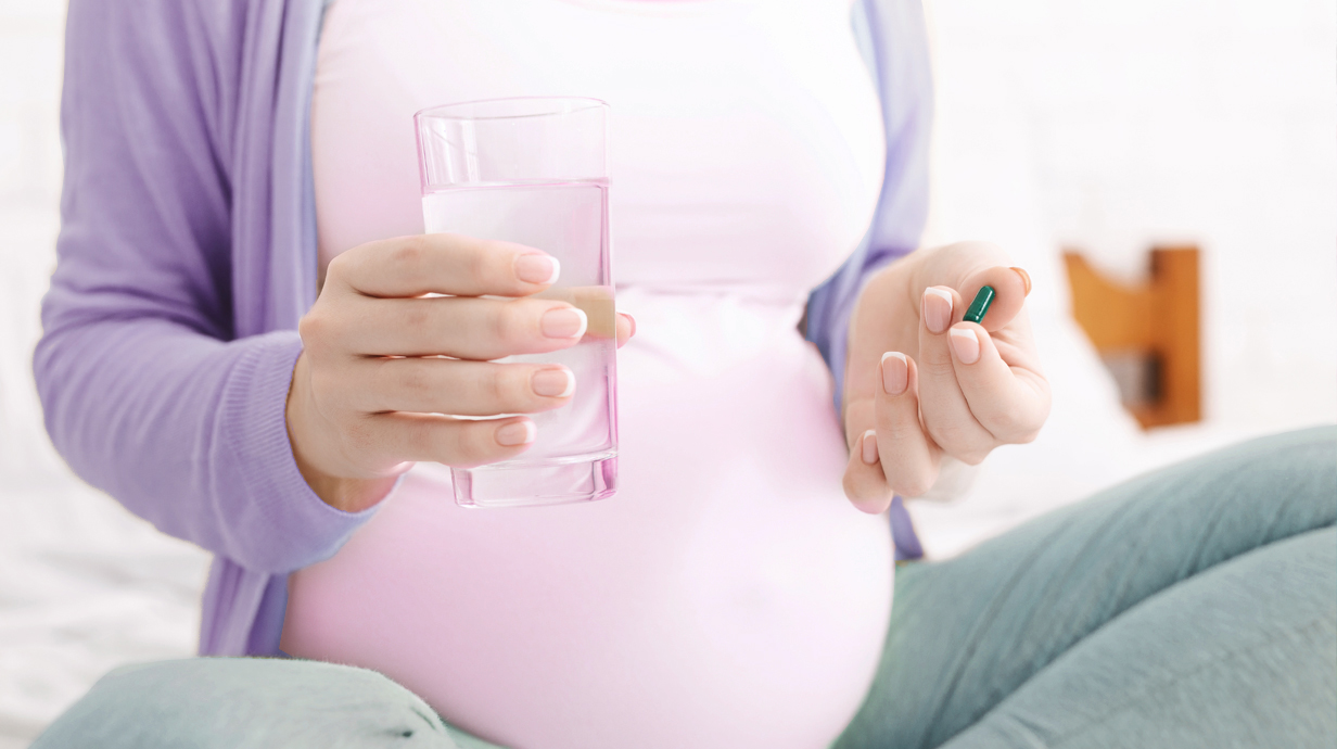 A pregnant woman holds a green pill and a glass of water.