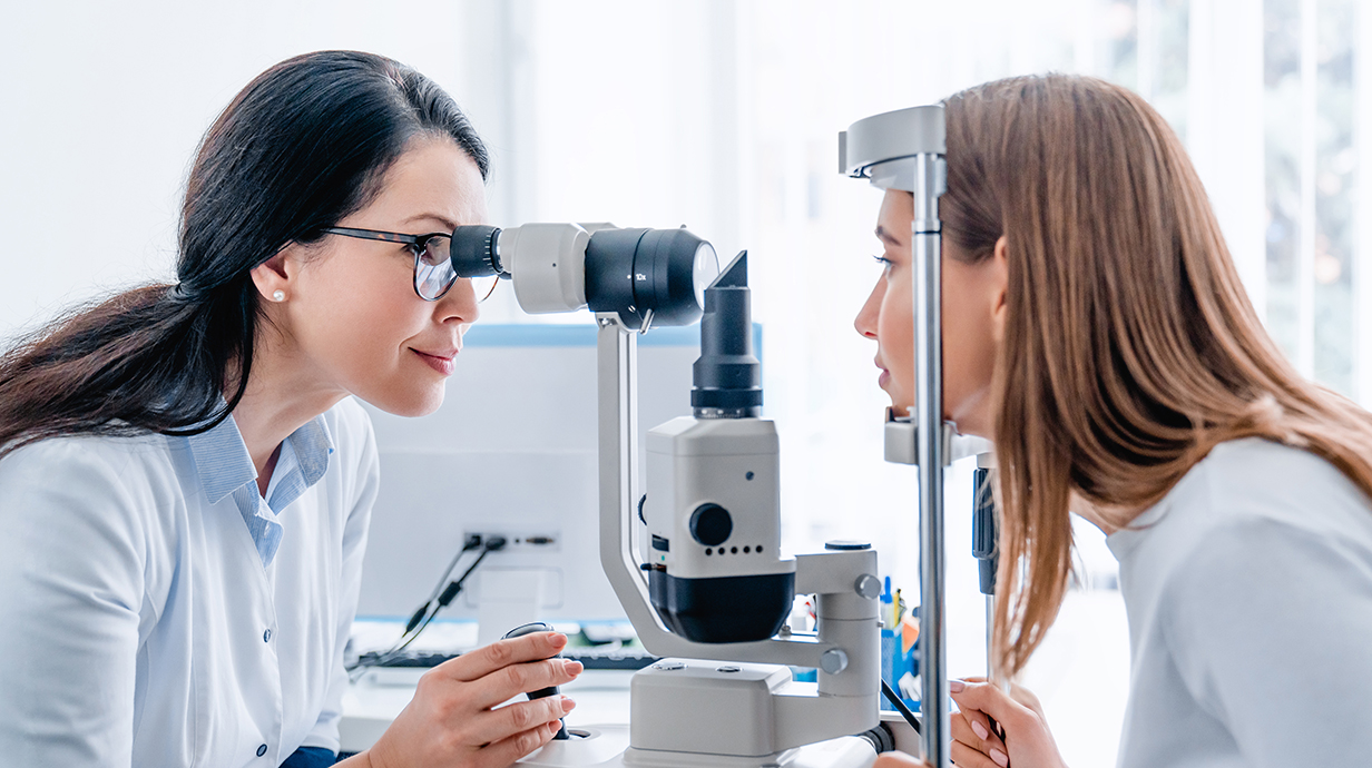 A woman looks through an ophthalmologist's equipment into another woman's eyes.