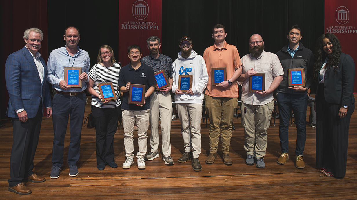 A man and a woman stand on a stage with eight young men and women holding award plaques.