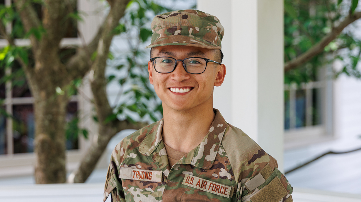 A young man wearing military fatigues stands on an outdoor porch.