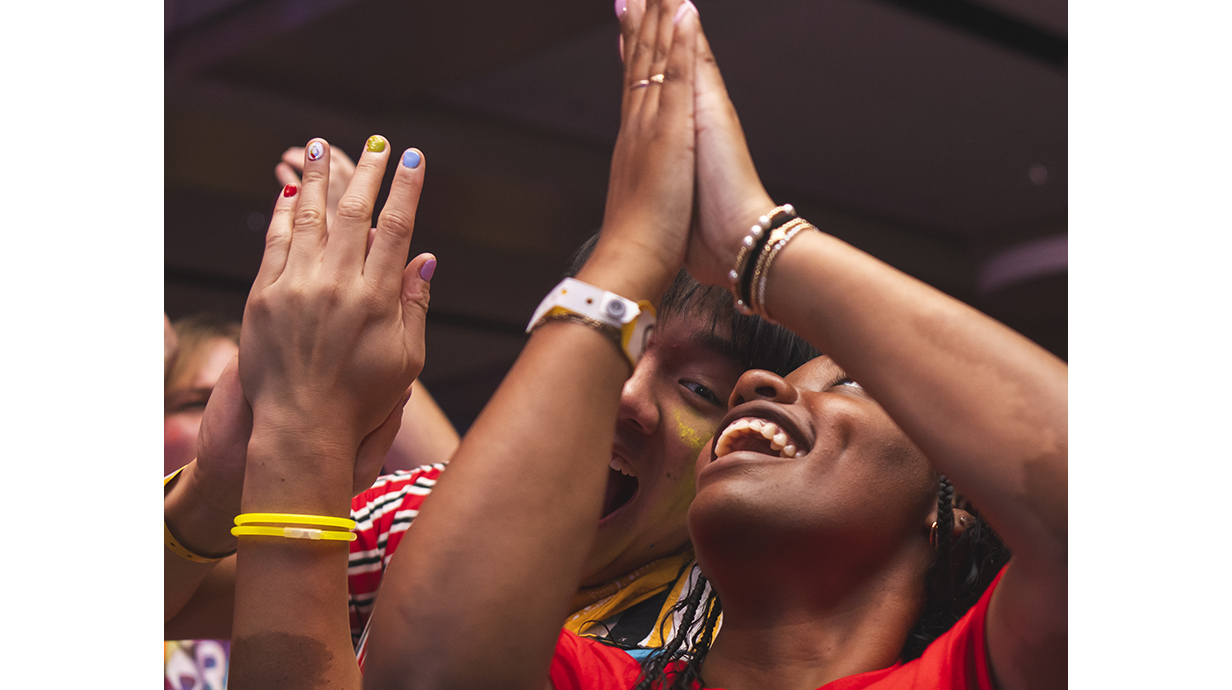 A young woman and young man stand together and clap as part of a crowd.