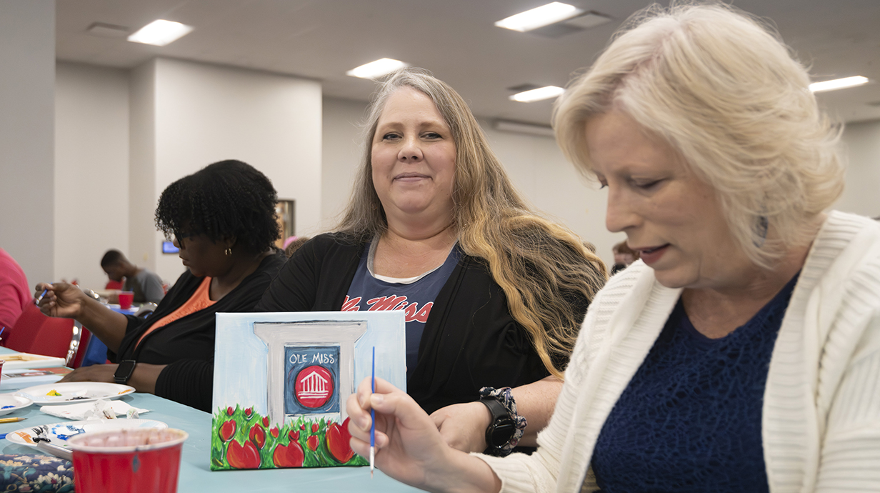 A woman shows off a completed painting while another woman works on a painting.