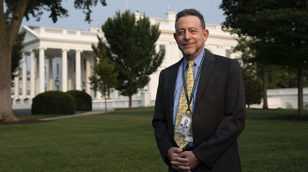 A man wearing a suit and name badges on a lanyard stands in front of the White House.