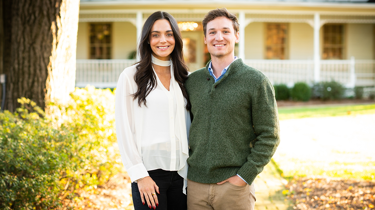 A young woman and man pose for a photo in front of an elegant white house.