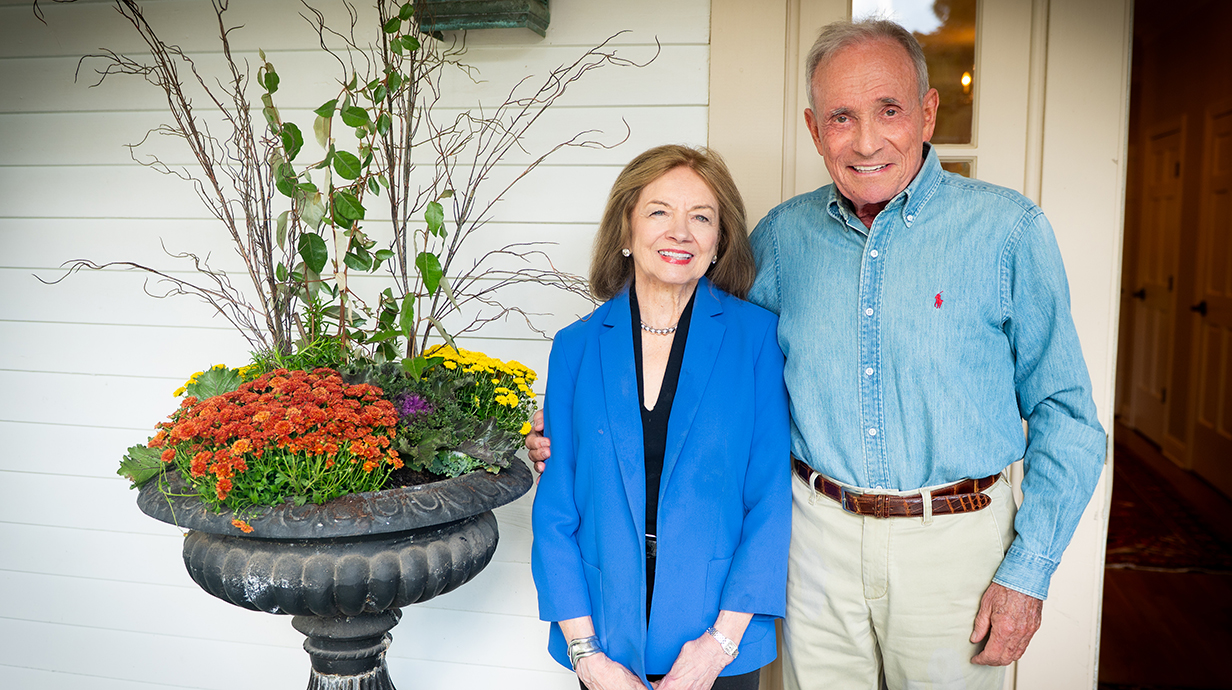 A man and woman stand on a porch next to a large potted plant.