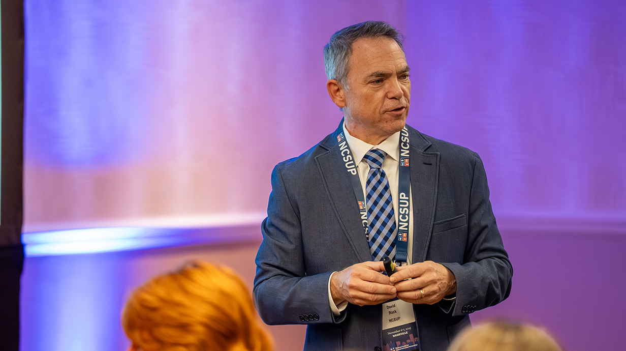 A man wearing a suit talks to a crowd from the front of a conference hall.
