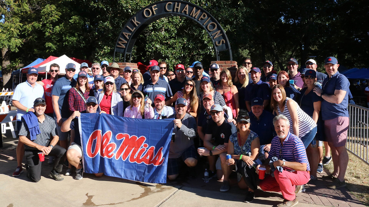 A large group of people hold an Ole Miss flag as they gather outdoors in front of the Walk of Champions arch.