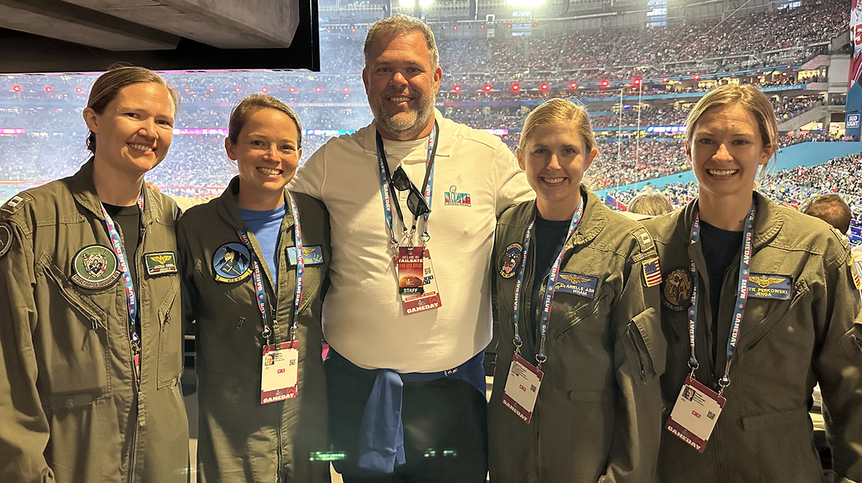 A man poses for a photo with four women, all wearing flight suits, in a sports arena.