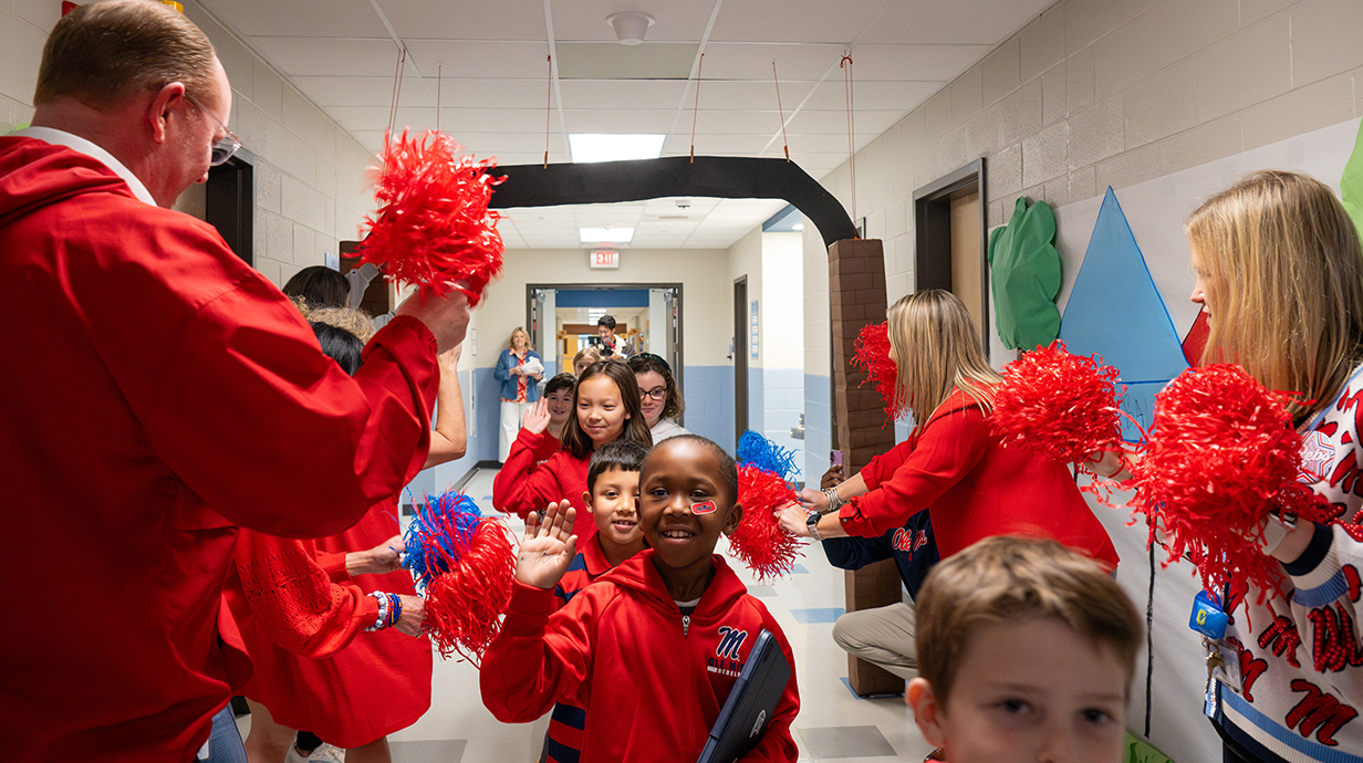 Elementary school students walk down a hallway while teachers greet them by cheering and shaking red pompoms.