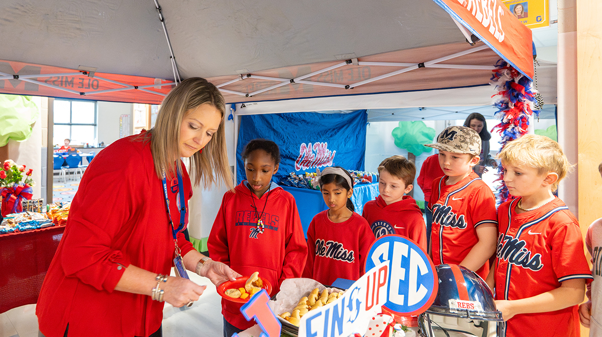 A woman serves snacks to several children, all wearing red Ole Miss gear, underneath a pop-up tent in a school hallway.