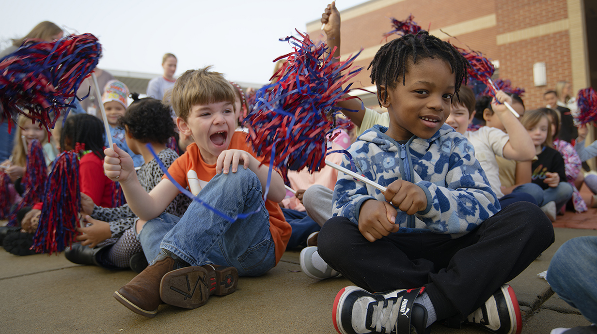 Children wave pompoms and cheer in front of a school.