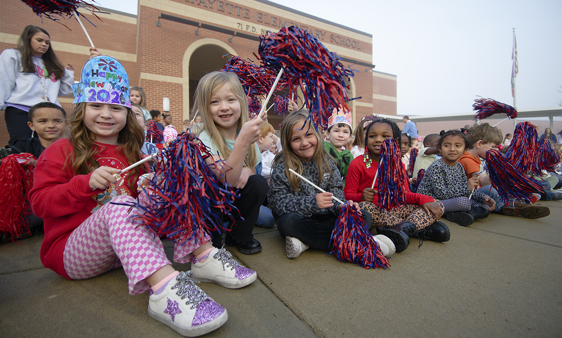 Children wave pompoms and cheer in front of a school.